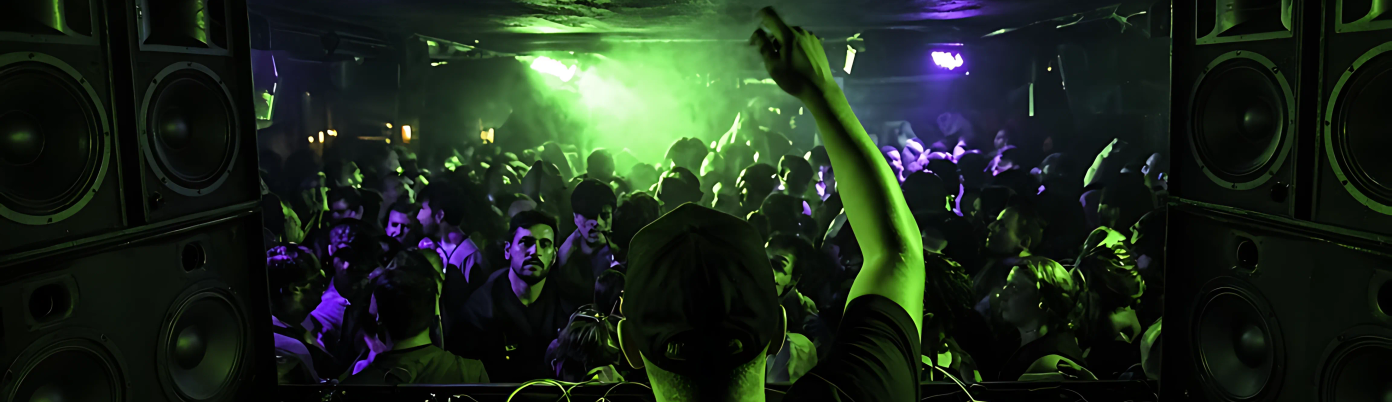 A DJ in the booth with a wall of speakers behind and a packed room.