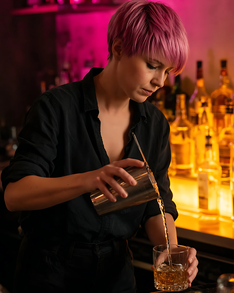 Tess Mallory pouring a drink behind a neon-lit bar.